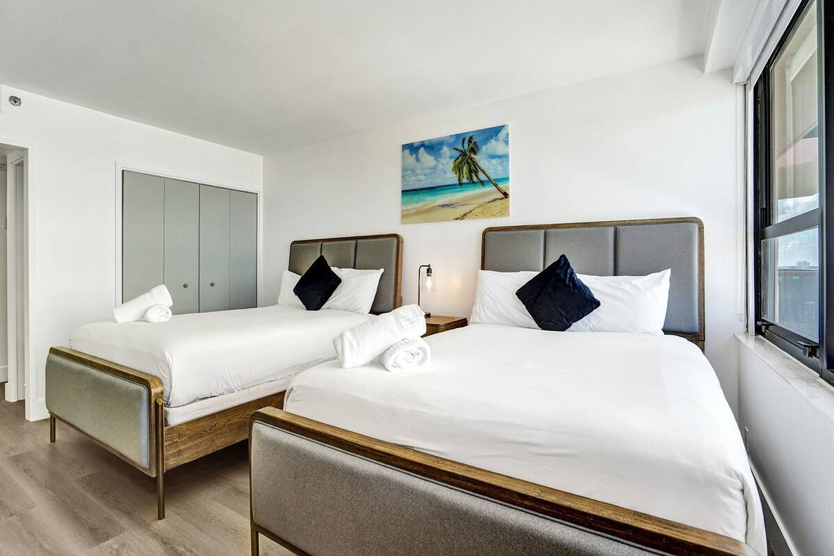 Quarto de hotel com duas camas de solteiro, uma janela grande e uma pintura de uma cena de praia.