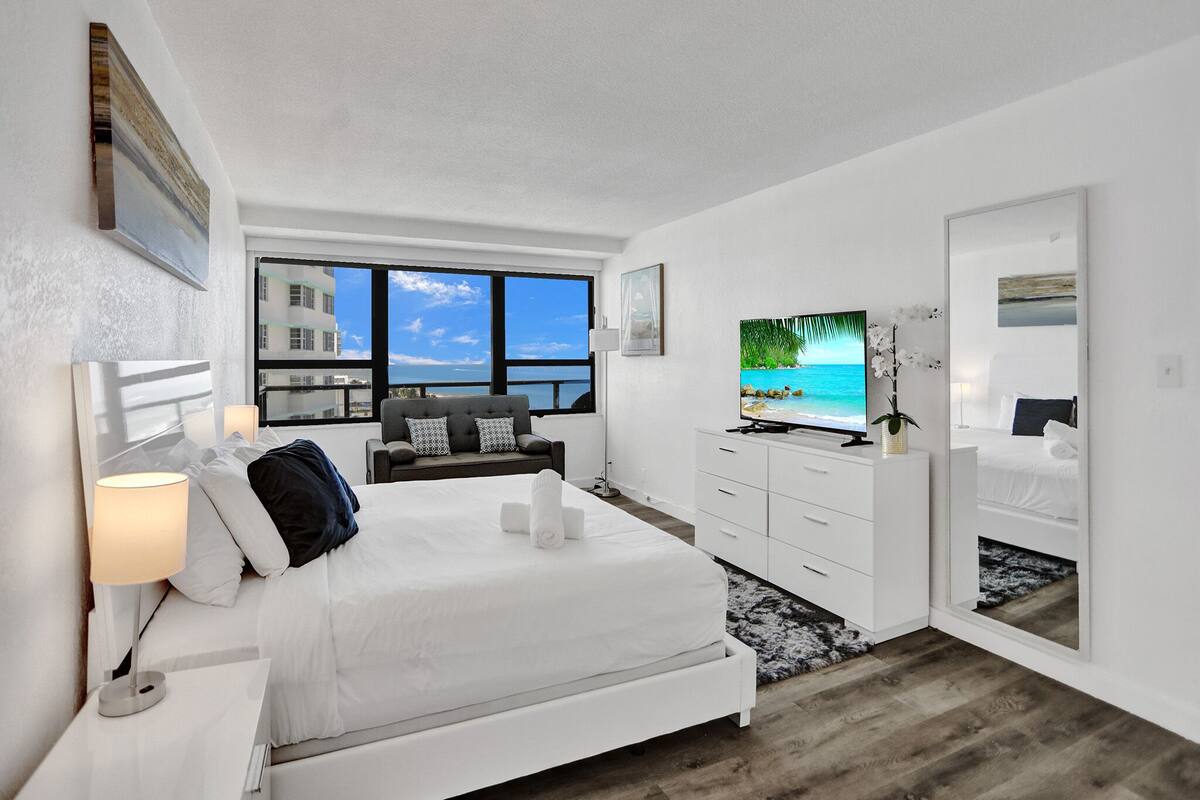 Quarto de hotel moderno com uma cama grande, televisão, sofá e vista para o mar pelas janelas.