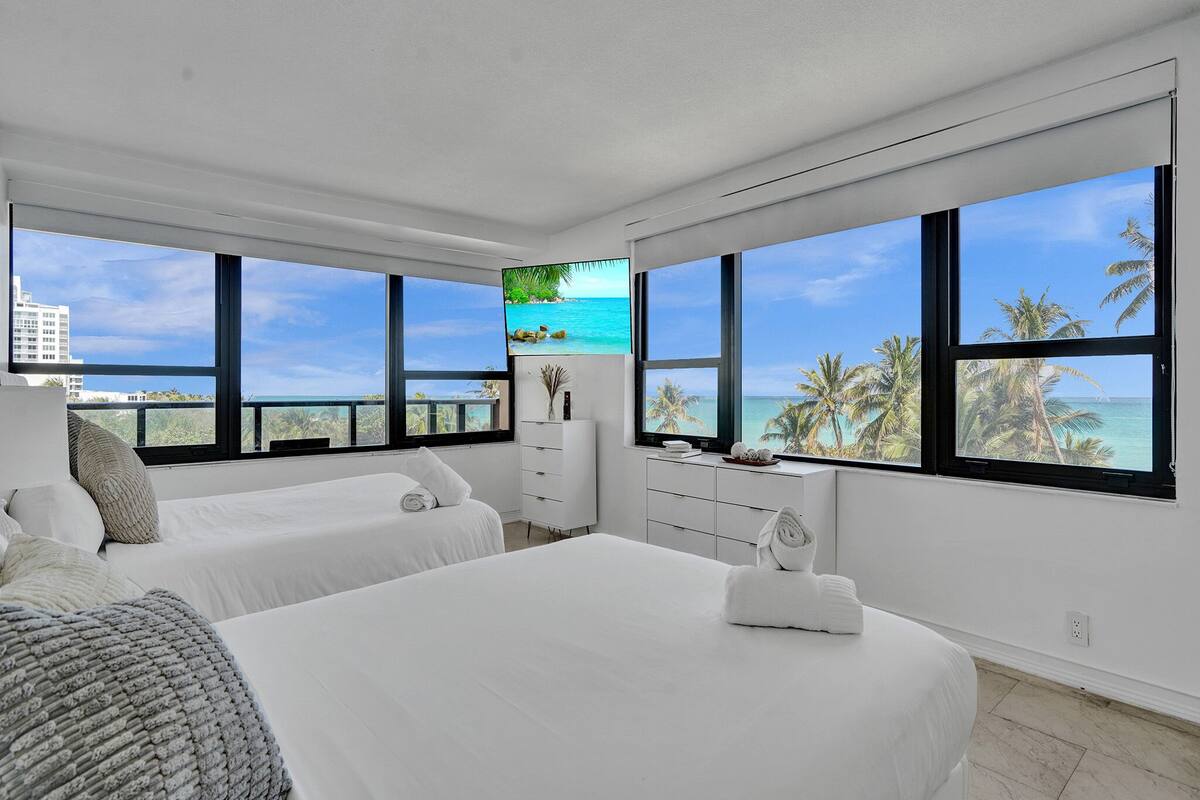 Quarto de hotel moderno com duas camas, uma janela grande com vista para o oceano, uma televisão de tela plana e uma cômoda branca.