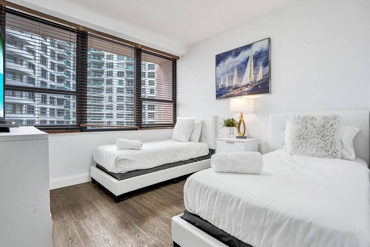Quarto de hotel moderno com duas camas, uma janela grande com cortinas e um quadro de barcos à vela, além de uma mesa de cabeceira com abajur.