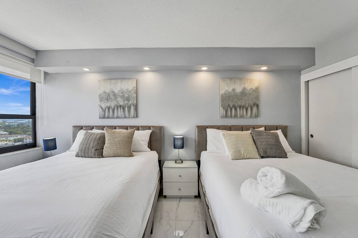 Quarto de hotel com duas camas, mesa de cabeceira com abajur e obras de arte nas paredes.