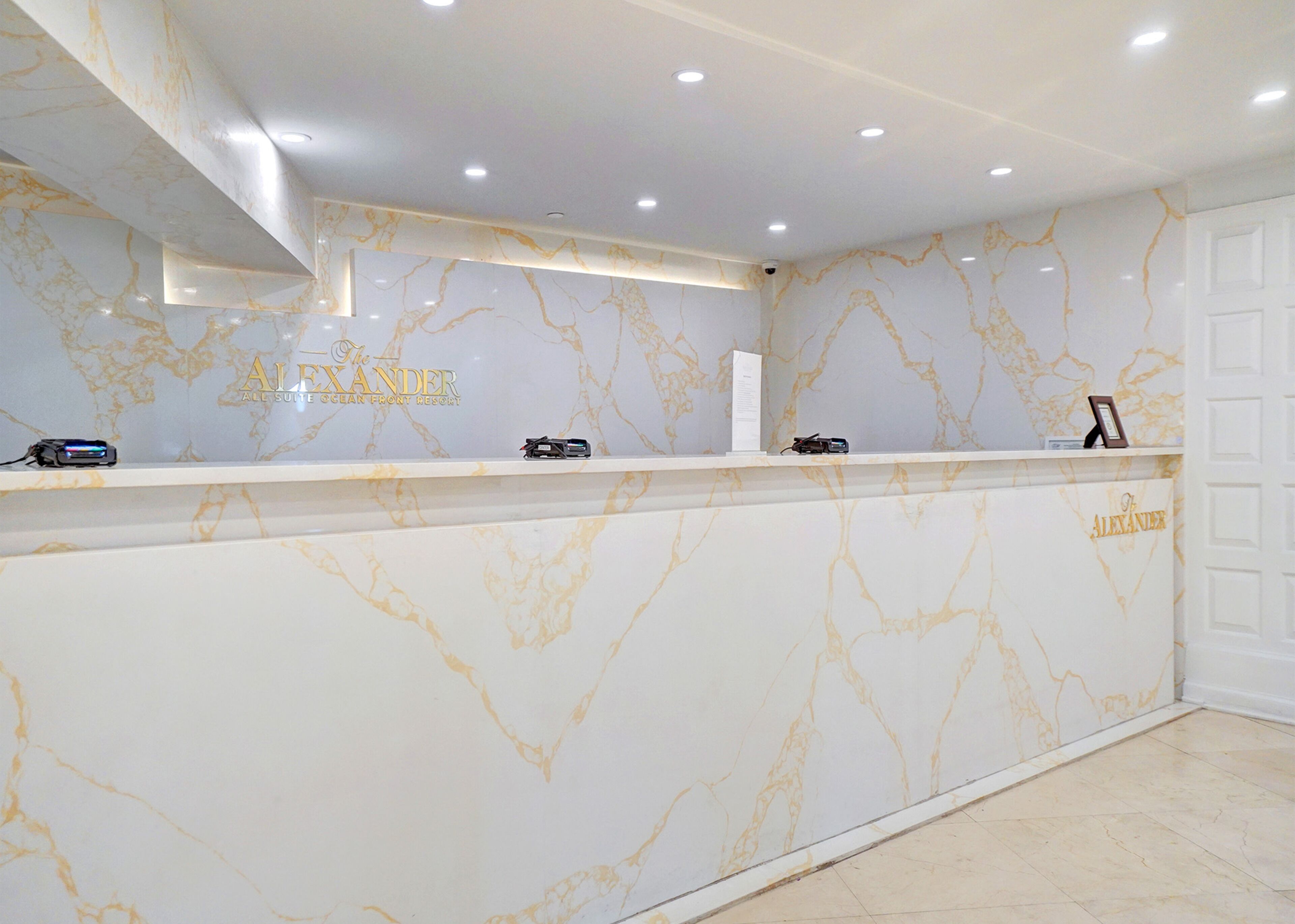 Un comptoir d’accueil au design en marbre, une enseigne indiquant « The Alexander All Suite Clean Front Resort », et une porte située à droite.