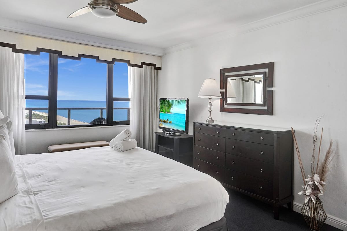 Quarto de hotel com cama, televisão, penteadeira, ventilador de teto e vista para o mar.