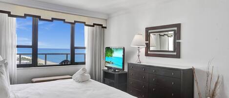 A hotel room with a bed, a television, a dresser, a ceiling fan, and a view of the sea.