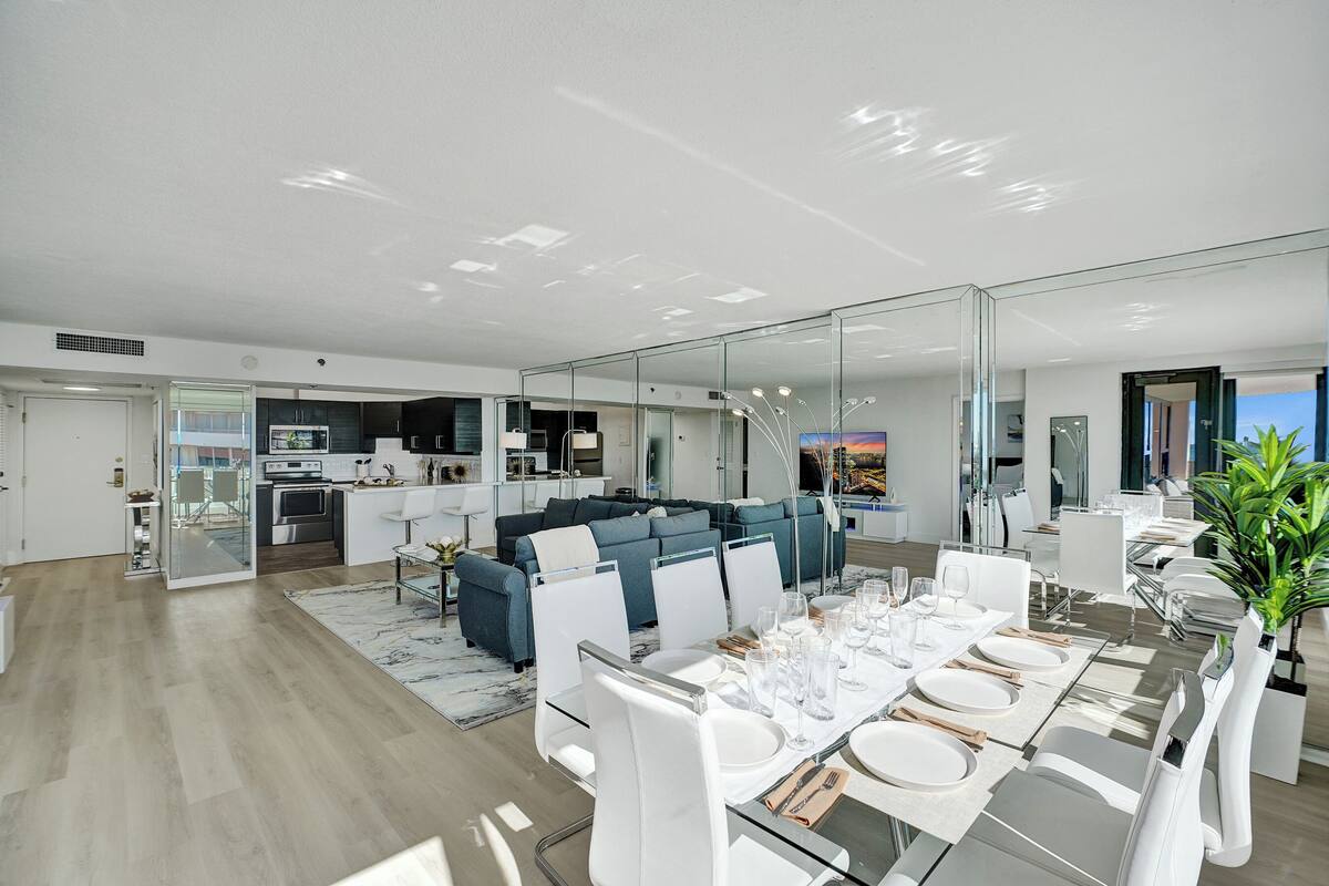 Uma zona de refeições moderna com mesa de vidro posta para seis, um sofá ao fundo e uma cozinha visível através de uma divisória de vidro.