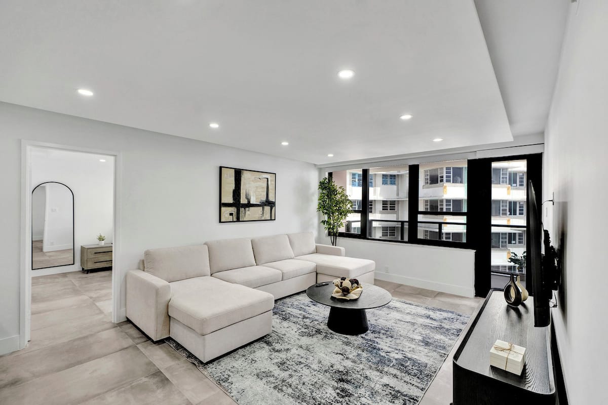 Uma sala de estar moderna com um sofá seccional, uma mesa de centro e uma grande janela com vista para edifícios.