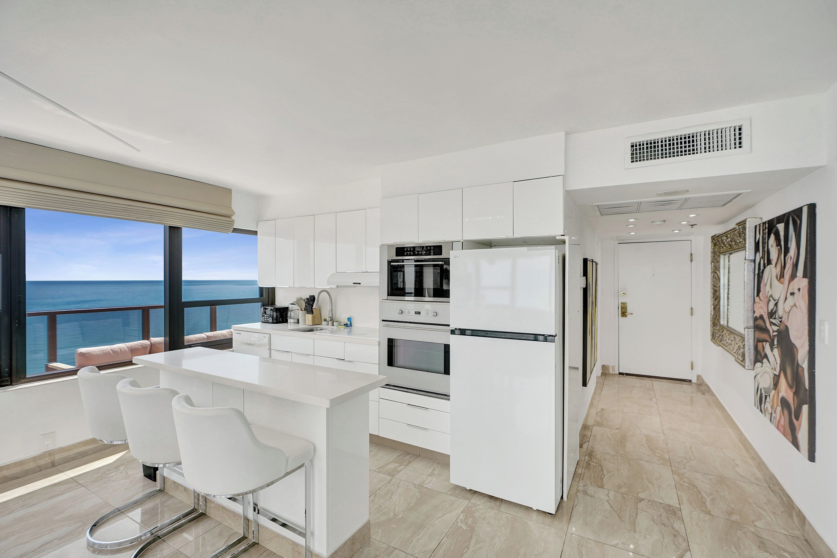 A modern kitchen with white cabinetry, a large island with bar stools, and a view of the sea.