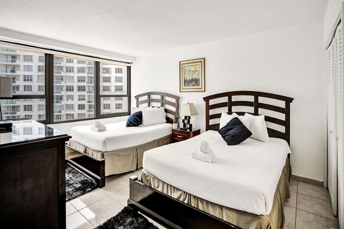 Quarto de hotel com duas camas, uma escrivaninha, uma cadeira, uma janela com vista para edifícios e um quadro na parede.