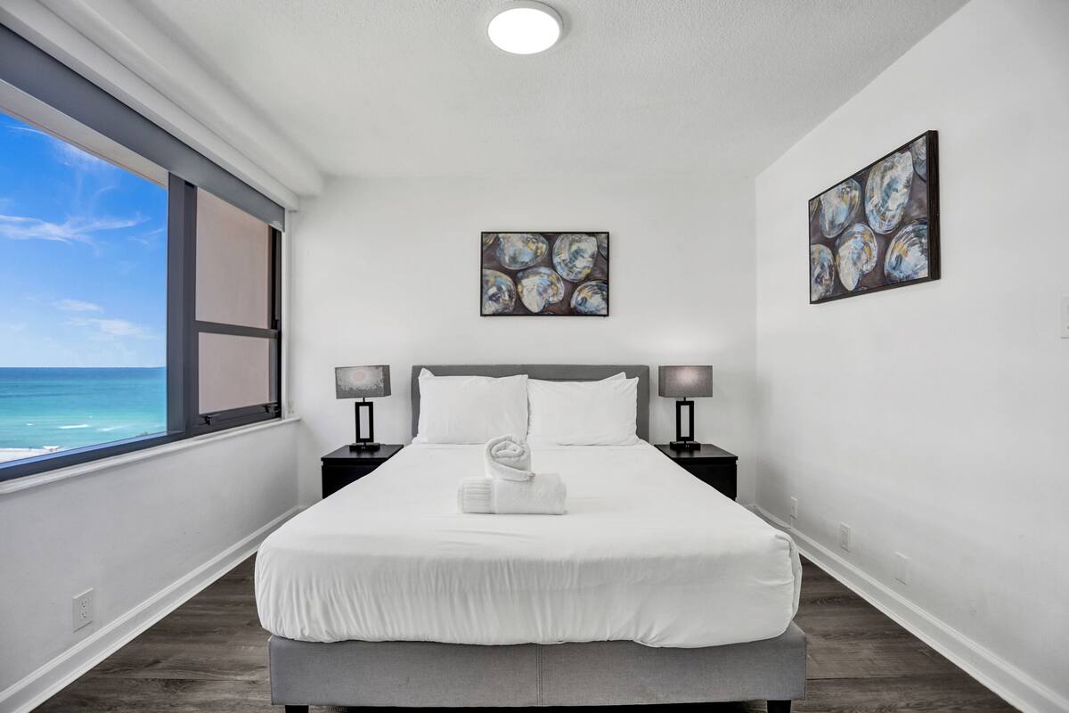Quarto de hotel com uma janela grande que oferece vista para o oceano, uma cama bem arrumada com toalha, duas luminárias de cabeceira e obras de arte nas paredes.