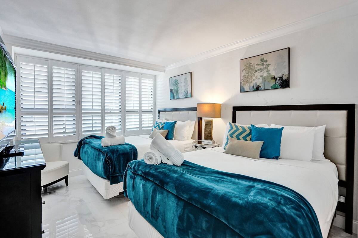 Quarto de hotel com duas camas, uma escrivaninha, uma cadeira, um abajur e obras de arte nas paredes.