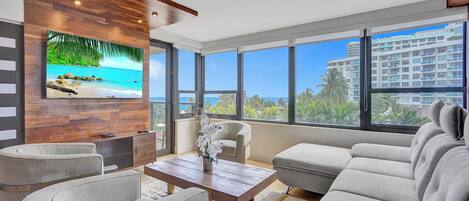 A modern living room with a large TV, comfortable seating, and a view of the ocean.