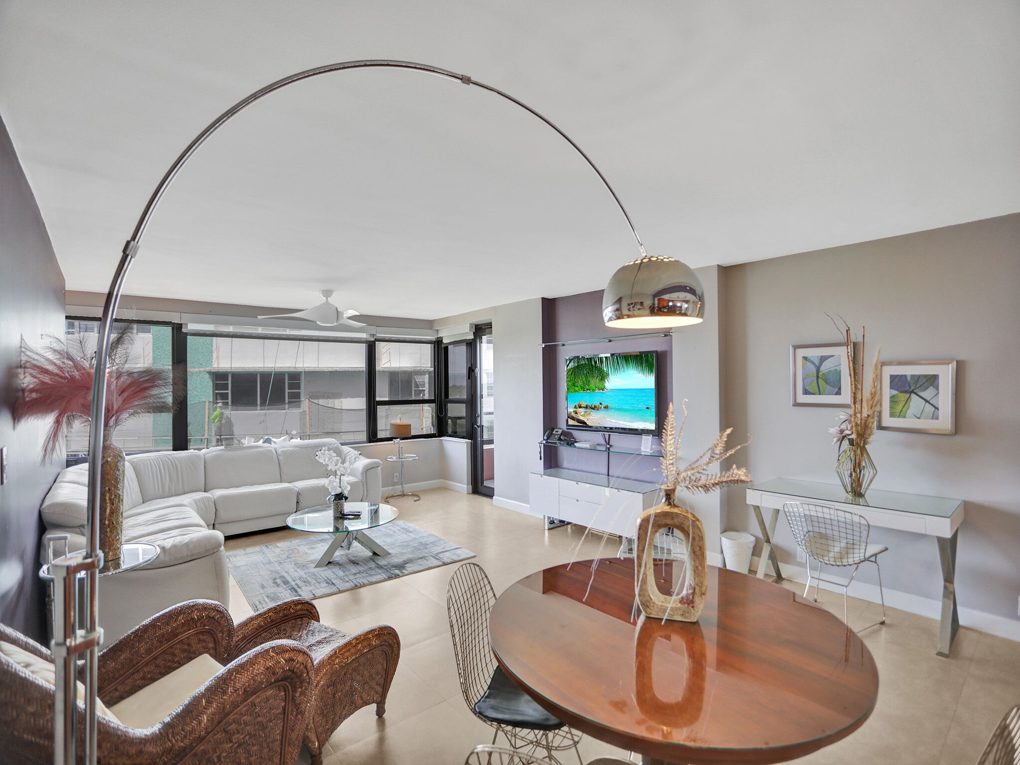 A modern living room with a white sofa, a round wooden dining table, wicker chairs, a glass coffee table, and a TV mounted on the wall.