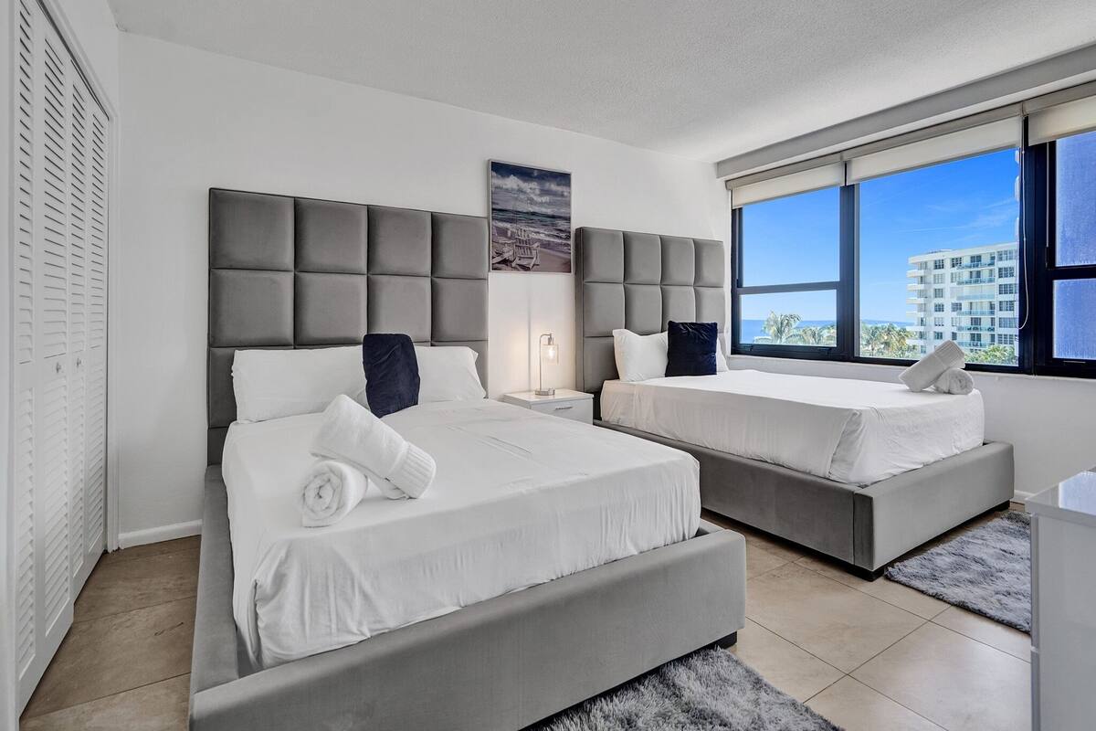 Quarto de hotel moderno com duas camas, uma janela ampla e vista para a cidade, e paleta de cores em tons de cinza.
