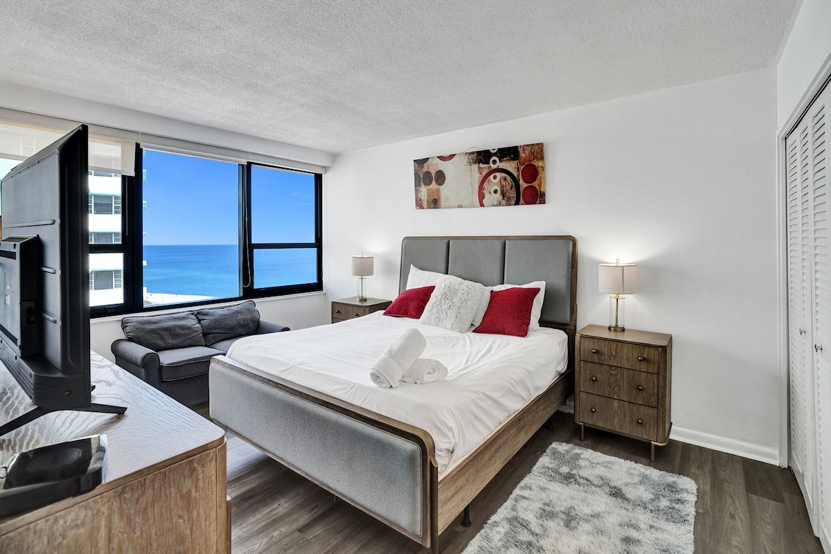 Quarto de hotel moderno com cama, escrivaninha, cadeira, televisão e vista para o oceano.
