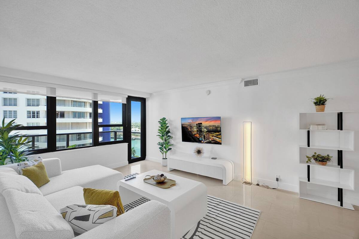 Uma sala de estar moderna com um sofá branco, uma mesa de centro com uma tigela decorativa, uma televisão de tela plana na parede e uma estante branca.