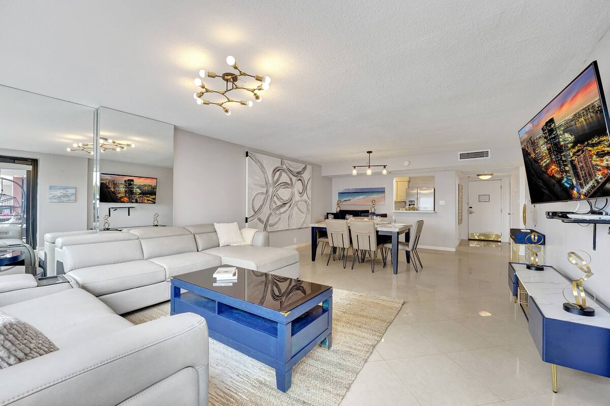 Uma sala de estar moderna com um sofá branco, uma mesa de centro azul e uma grande televisão de tela plana.
