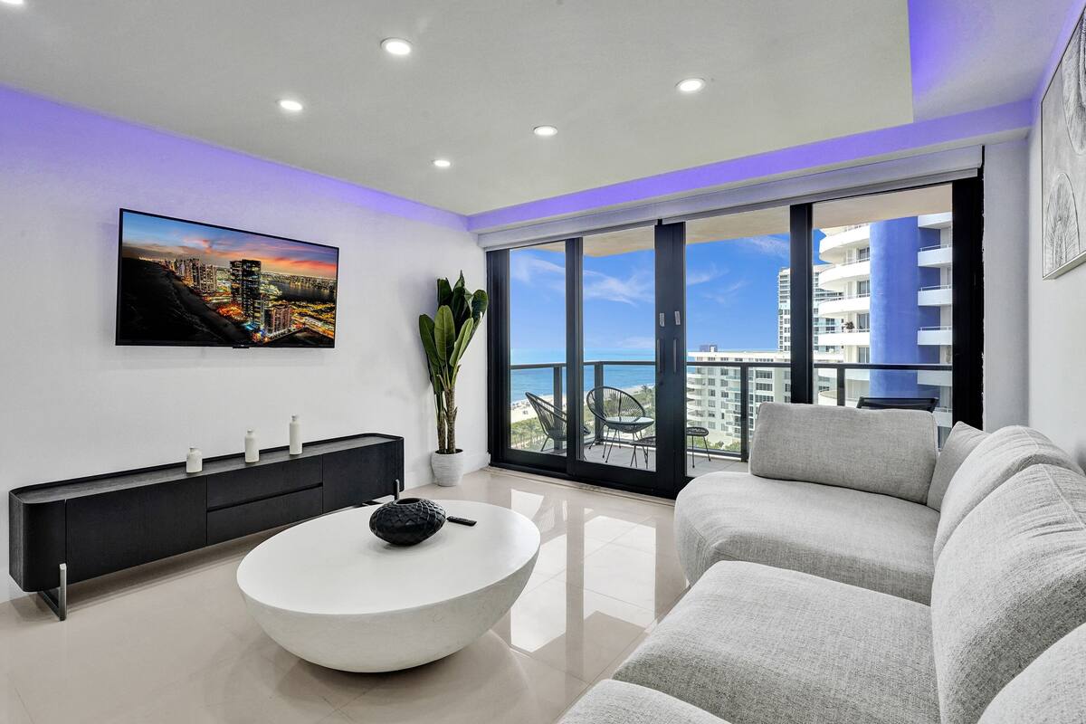 Uma sala de estar moderna com uma televisão de tela plana grande, uma mesa de centro redonda, um sofá cinza e uma varanda com vista para a cidade.