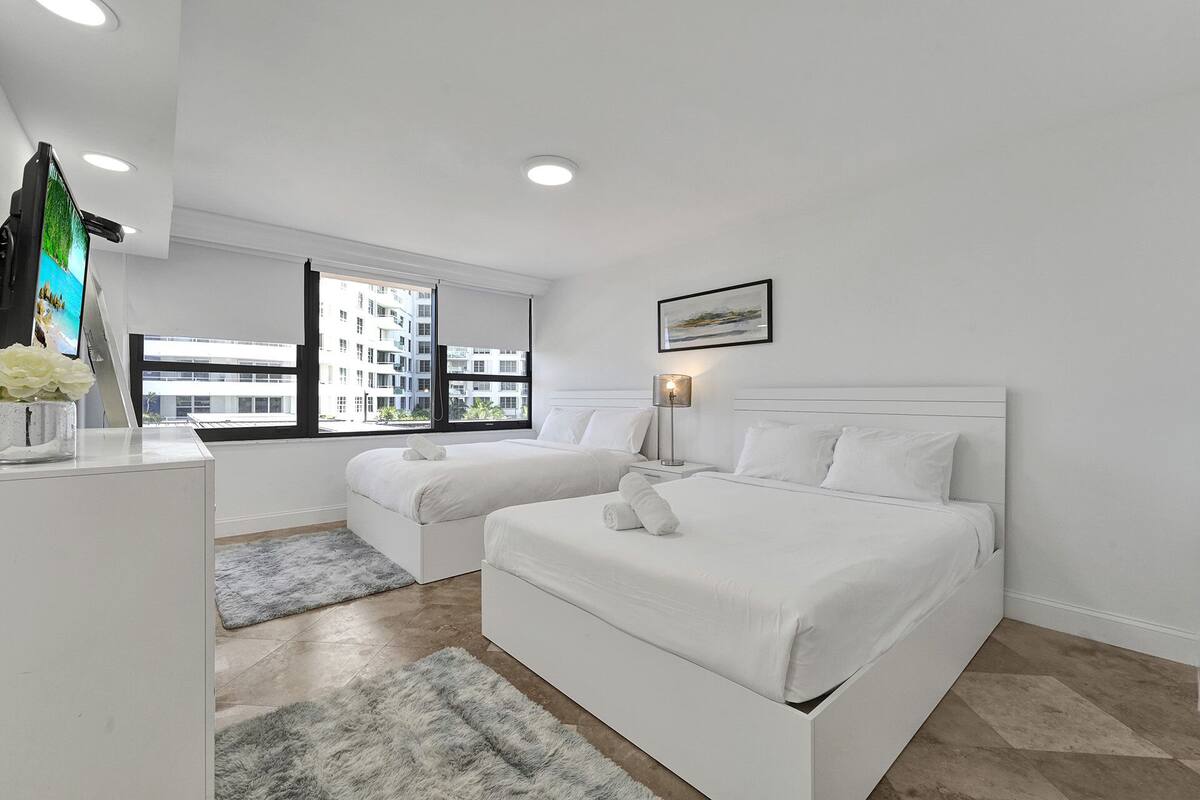 Quarto de hotel moderno com duas camas, uma escrivaninha e uma janela grande com vista para a cidade.