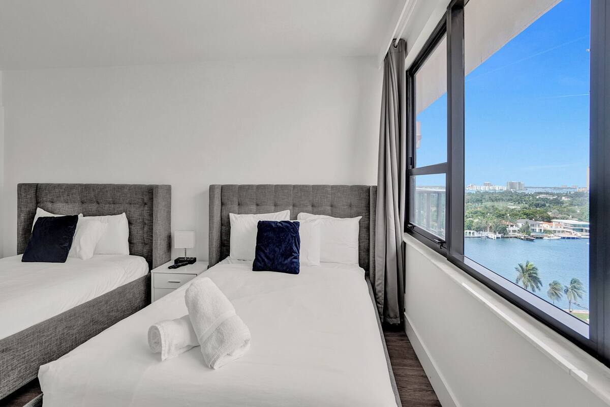 Quarto de hotel com duas camas, uma janela grande com vista para a cidade e uma cama bem arrumada com toalha branca.