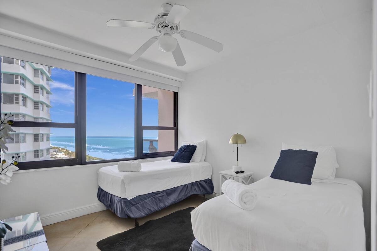 Quarto de hotel com duas camas, um ventilador de teto, mesa de cabeceira com abajur e vista para o oceano.