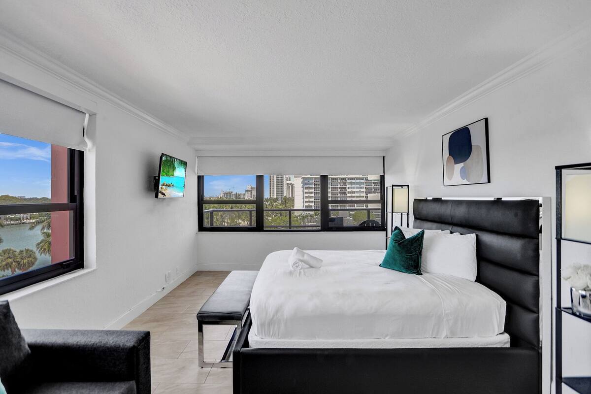 Quarto de hotel moderno com uma cama grande, um banco, uma TV e vista para a cidade pelas janelas.