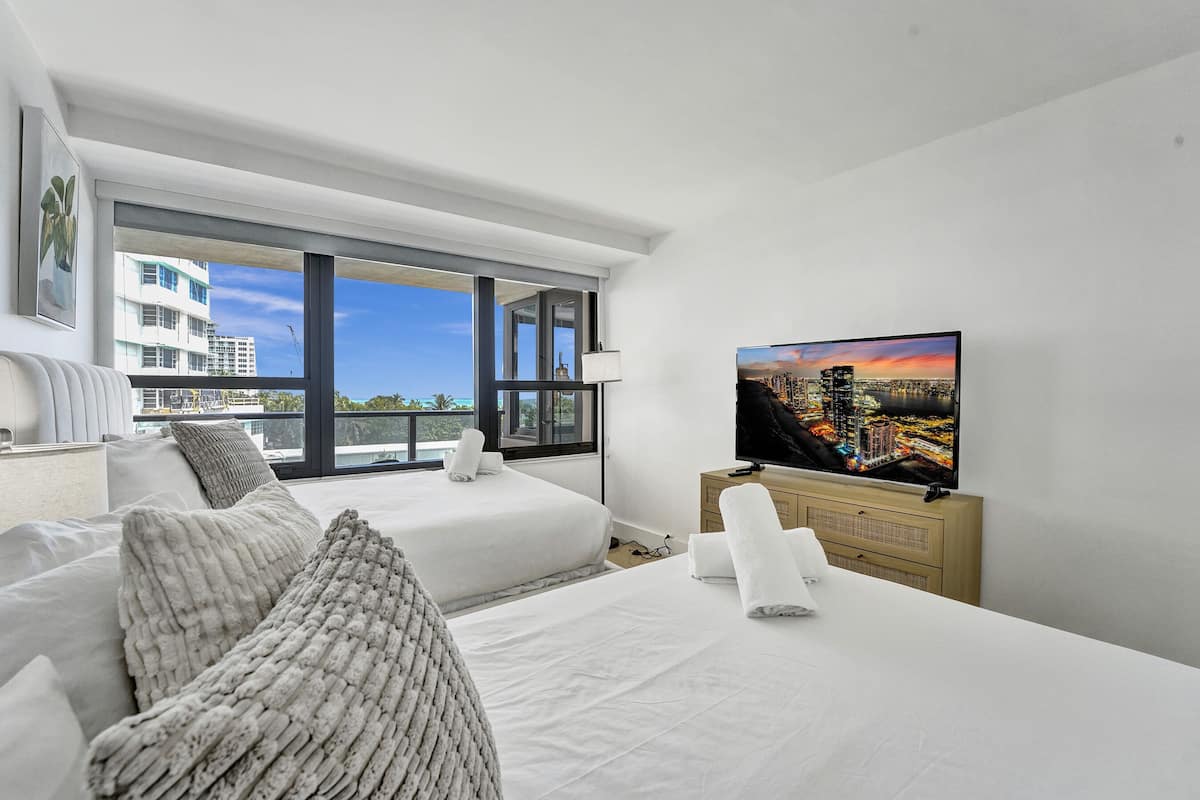 Quarto de hotel moderno com uma cama grande, televisão e varanda com vista para a cidade.