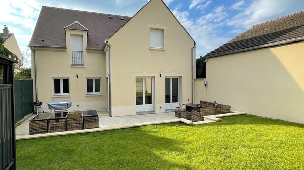 A two-story house with a flat roof, a covered patio with outdoor furniture, and a well-maintained lawn. - Ecrin de charme (Compiègne)