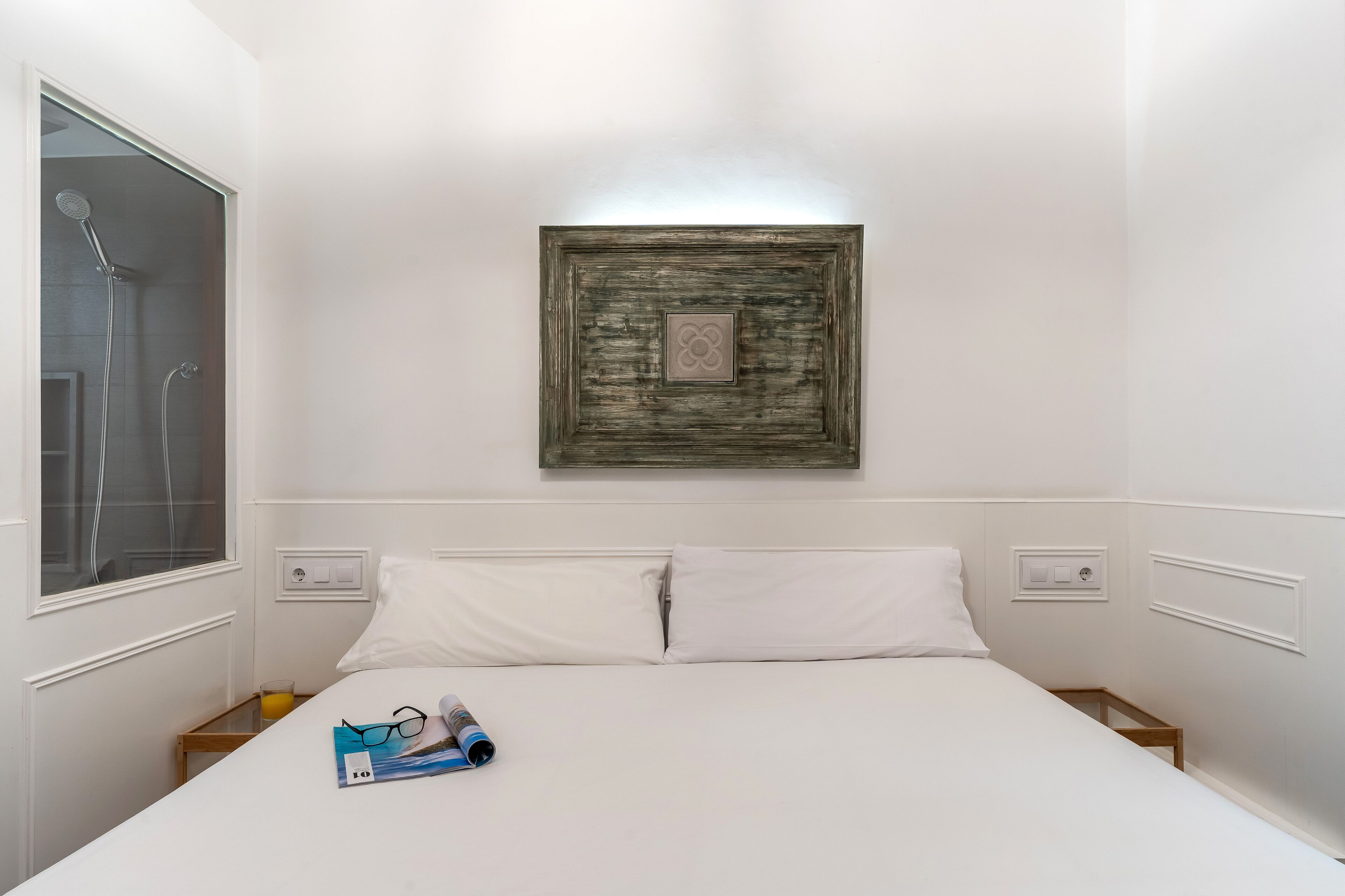 A neatly made bed with white linens, a small bedside table with a glass of orange juice, and a framed picture on the wall.