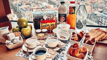 Una mesa de desayuno con diversos alimentos y bebidas, y una vista urbana al fondo.