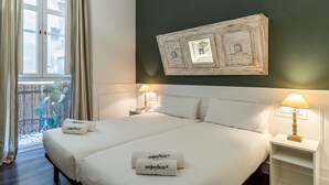 A double bed with white linens and two towels with "enjoyben" text, a nightstand with a lamp, a wall-mounted artwork, and a window with curtains. - Enjoybcn Tallers Apartment (Barcelona)