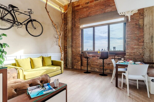 A cozy living area with a yellow sofa, a brown leather couch, and a dining table with chairs. A bicycle is mounted on the wall, and a large window offers a view of the city. - Enjoybcn Plaza Catalunya Apartment (Barcelona)