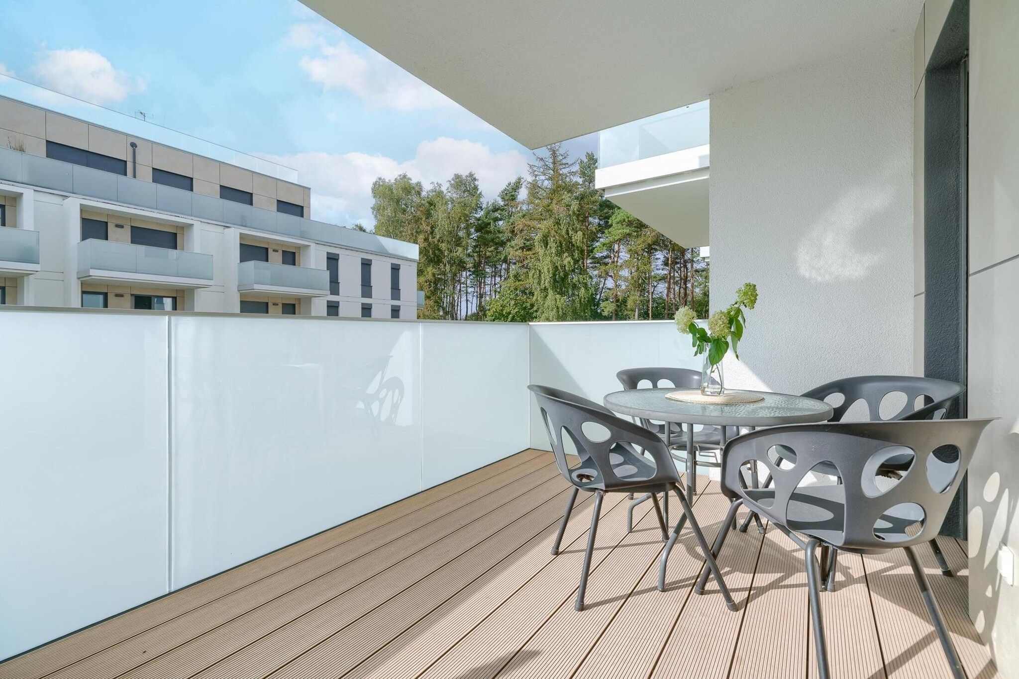 A modern outdoor patio with a round table and chairs, a potted plant, and a view of a building with balconies and trees.