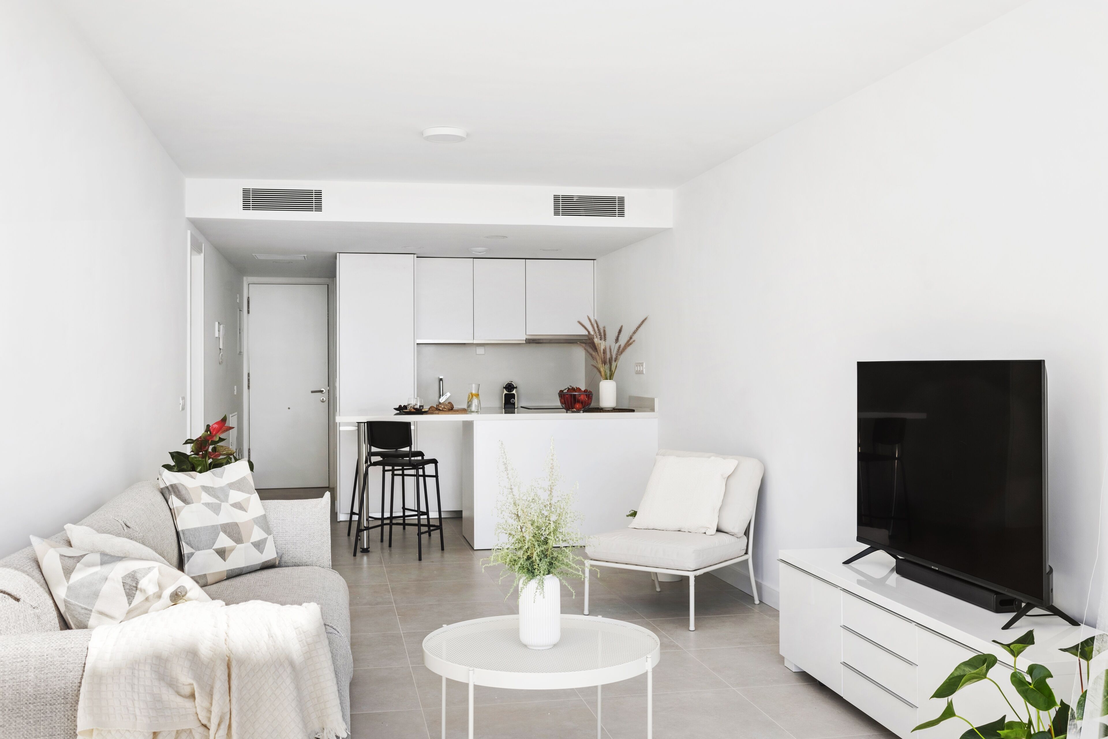 A modern living room with a sofa, coffee table, and TV. A kitchen is visible in the background with a dining area.