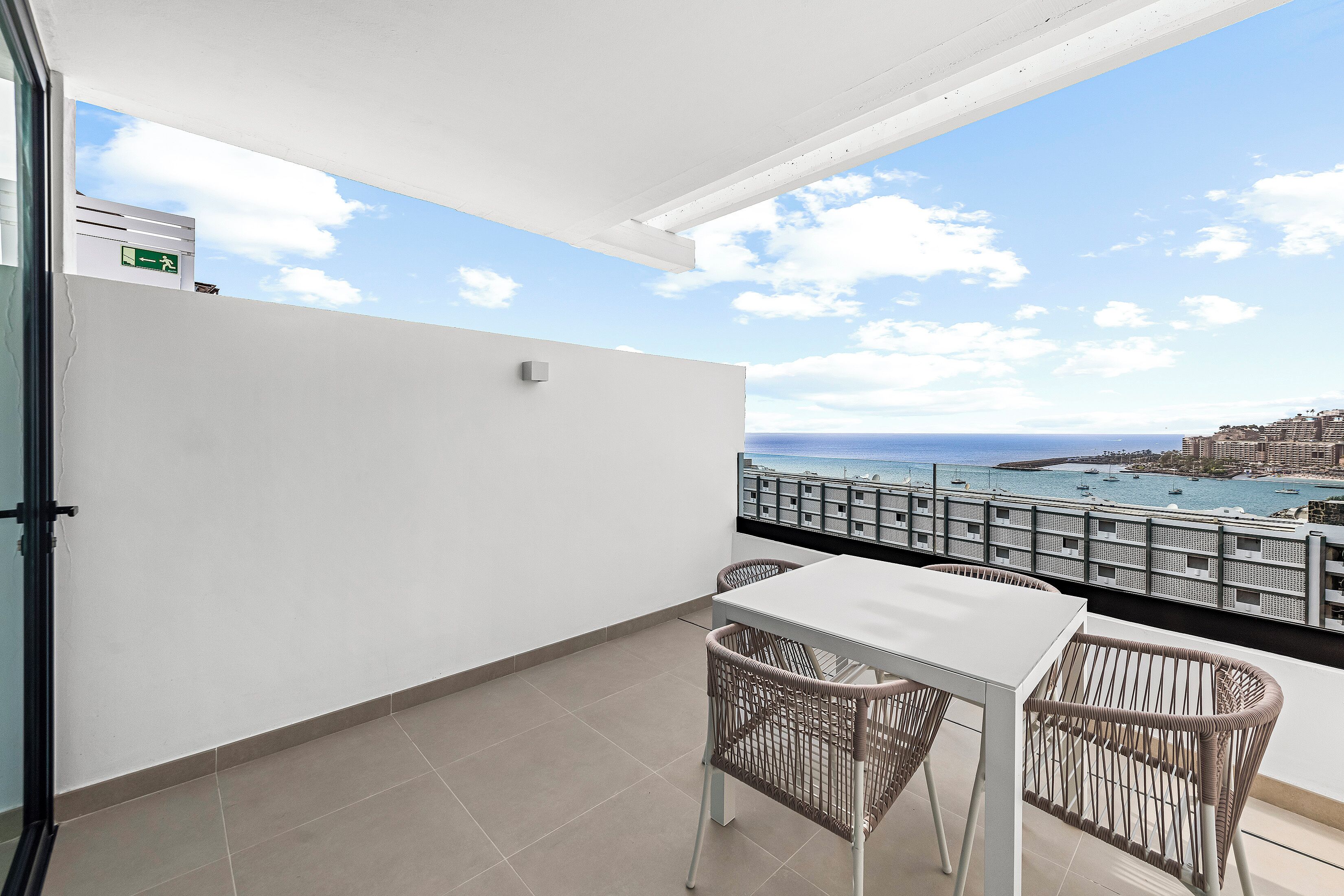A balcony with a white table and wicker chairs, overlooking a coastal view with boats and buildings.
