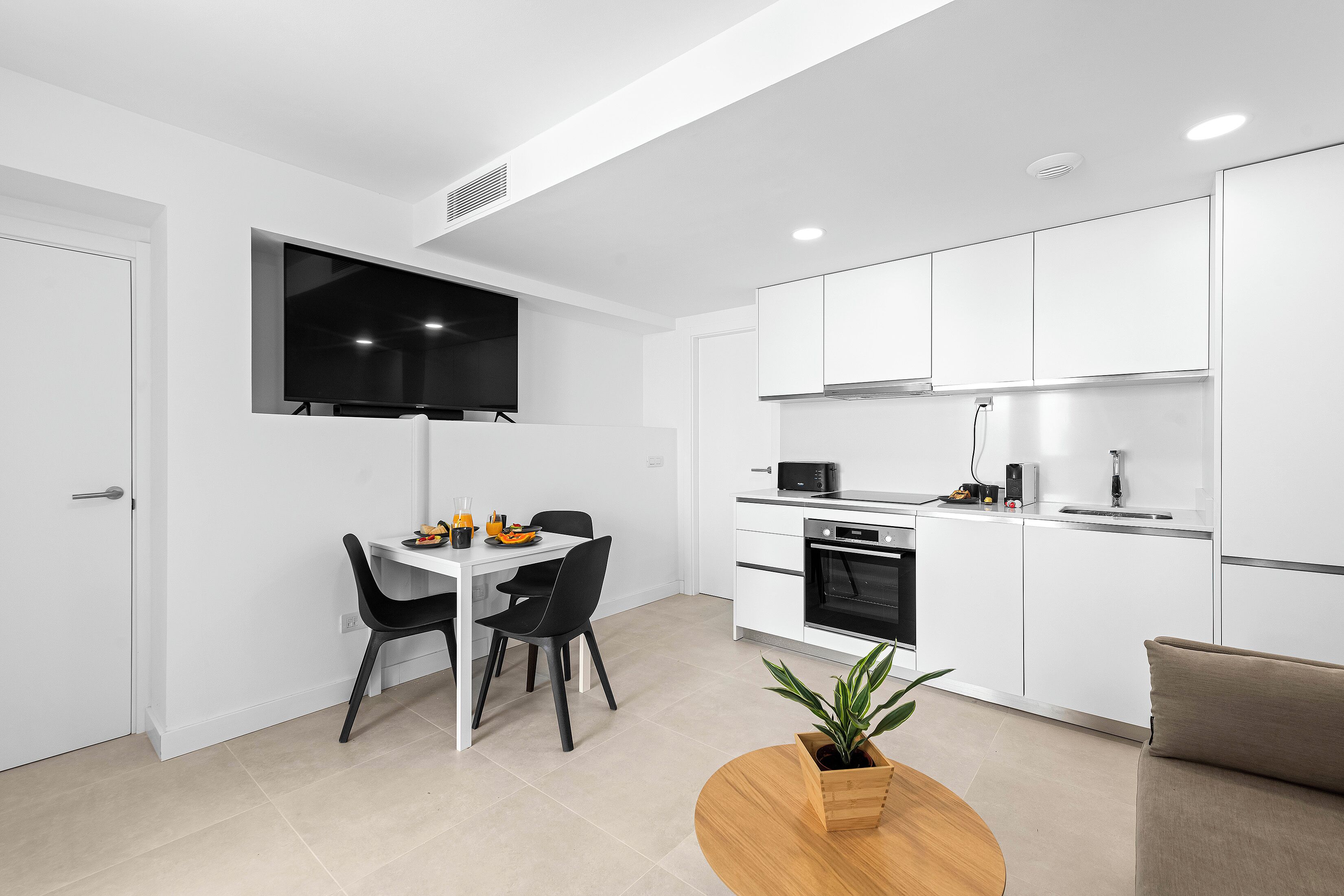 Una cocina moderna con armarios blancos, microondas y fregadero. Hay una zona de comedor con una mesa y sillas, y una planta sobre una mesa de madera.