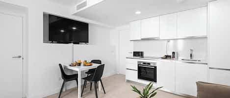 A modern kitchen with white cabinets, a microwave, and a sink. There is a dining area with a table and chairs, and a plant on a wooden table.