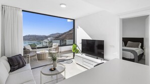 A modern living room with a large TV, a sofa, a dining table, and a balcony with a view of the sea and hills. - Palmera Sea View (Mogan)