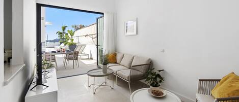 A modern living room with a sofa, two chairs, a coffee table, and a TV. There is a balcony with a table and chairs, and a view of the city.