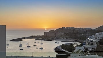 Paesaggio urbano costiero al tramonto, con edifici, un porto turistico e imbarcazioni.