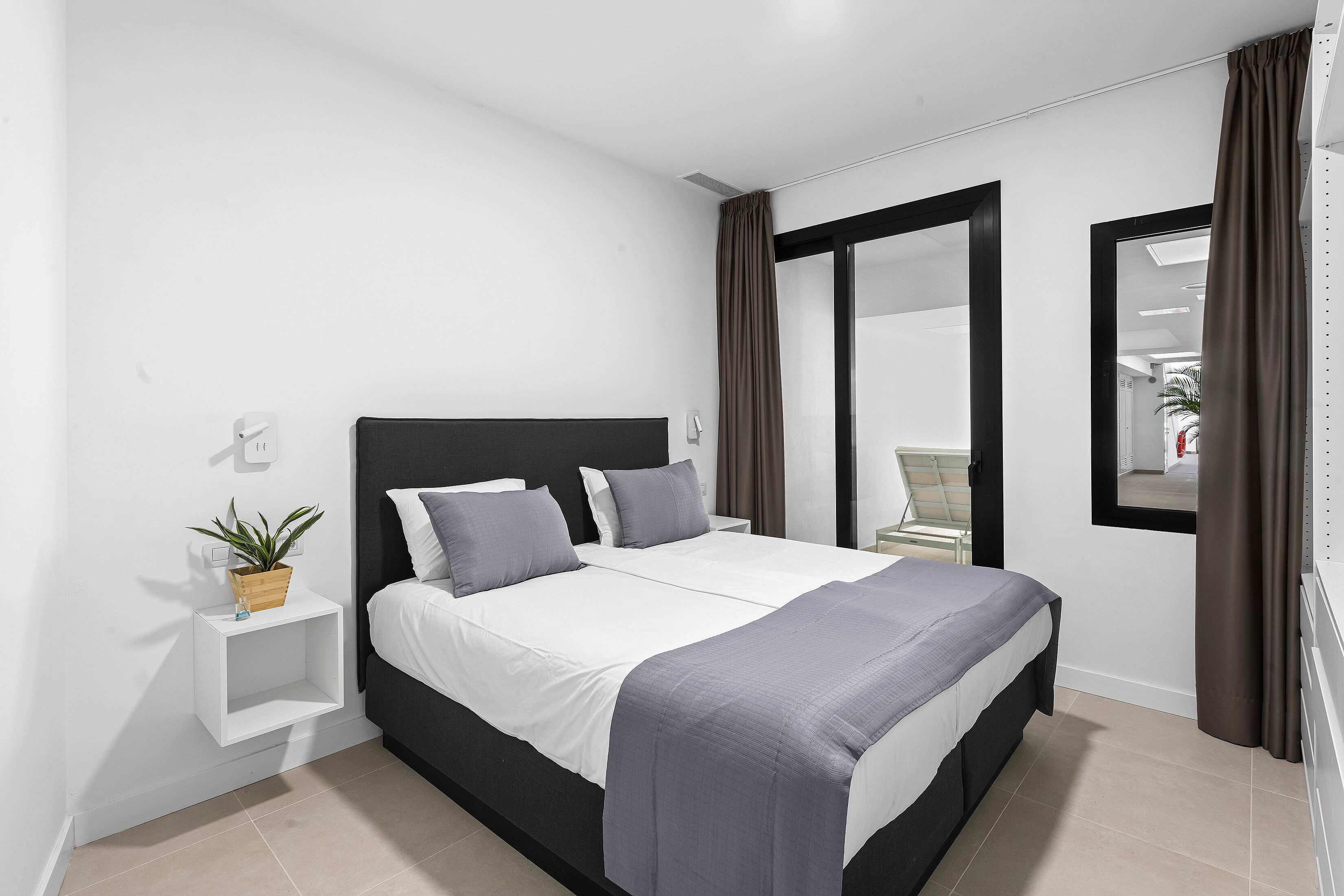 Un dormitorio moderno con cabecera negra, ropa de cama blanca y una mesita de noche con una planta.