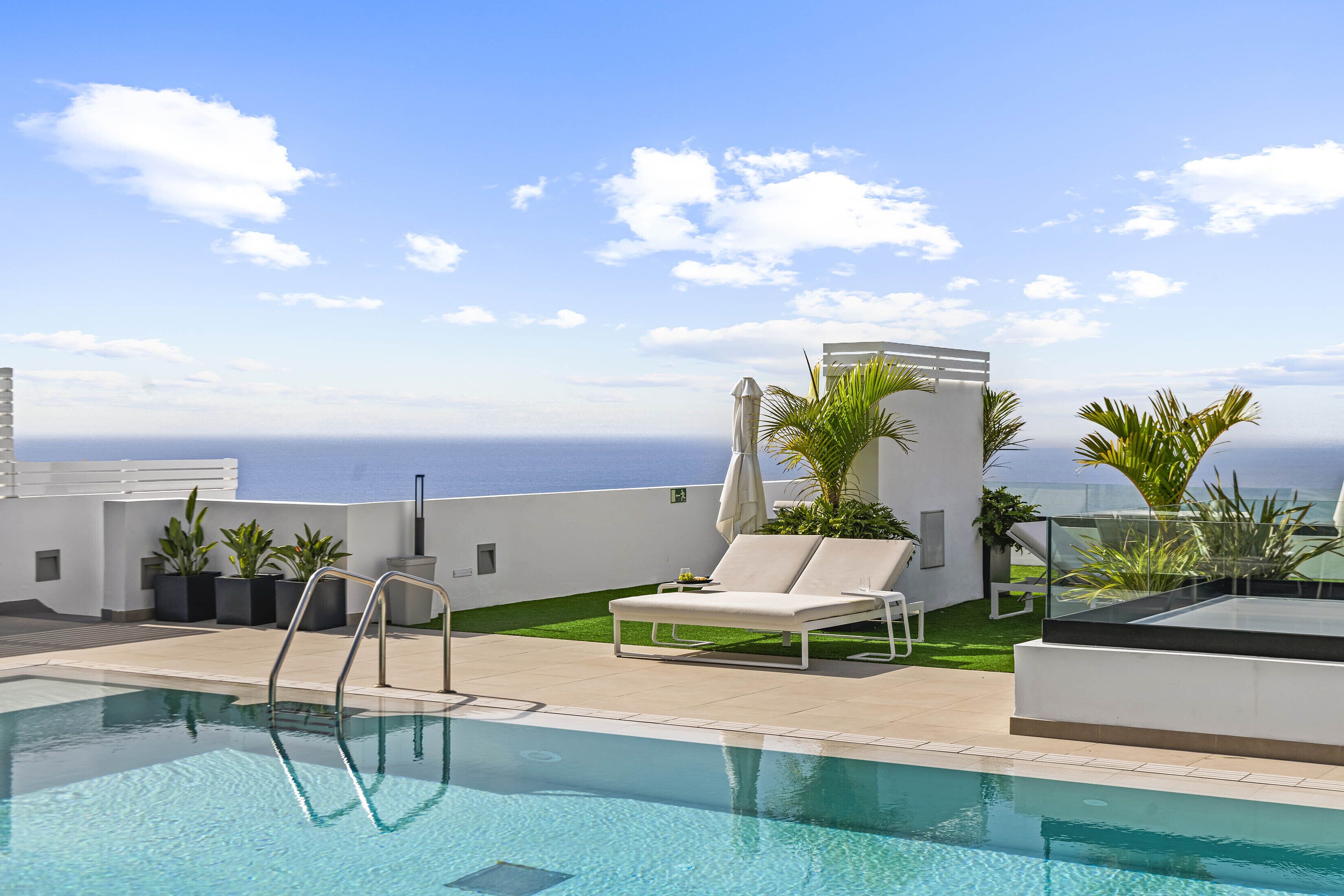 Un espace piscine aménagé sur le toit, offrant une vue dégagée sur l’océan, avec des chaises longues et des plantes en pot.