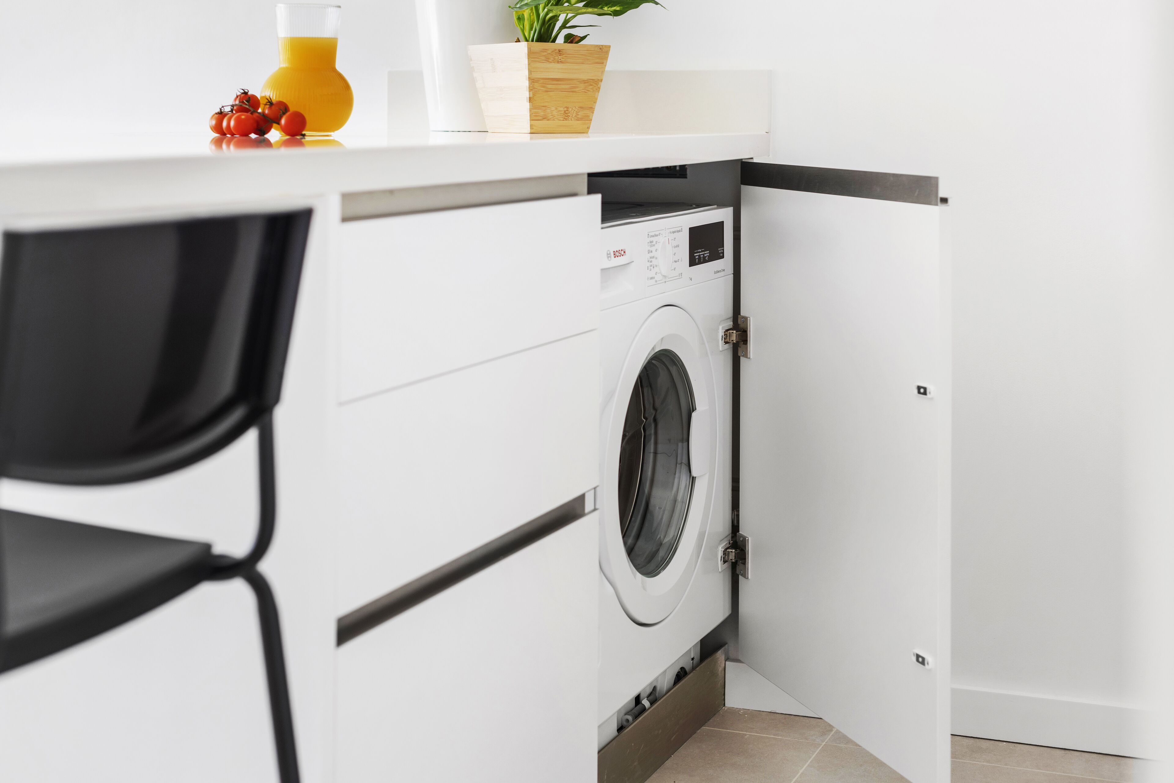 A compact kitchen with a built-in washing machine, a chair, and a small table with a plant and a bottle.