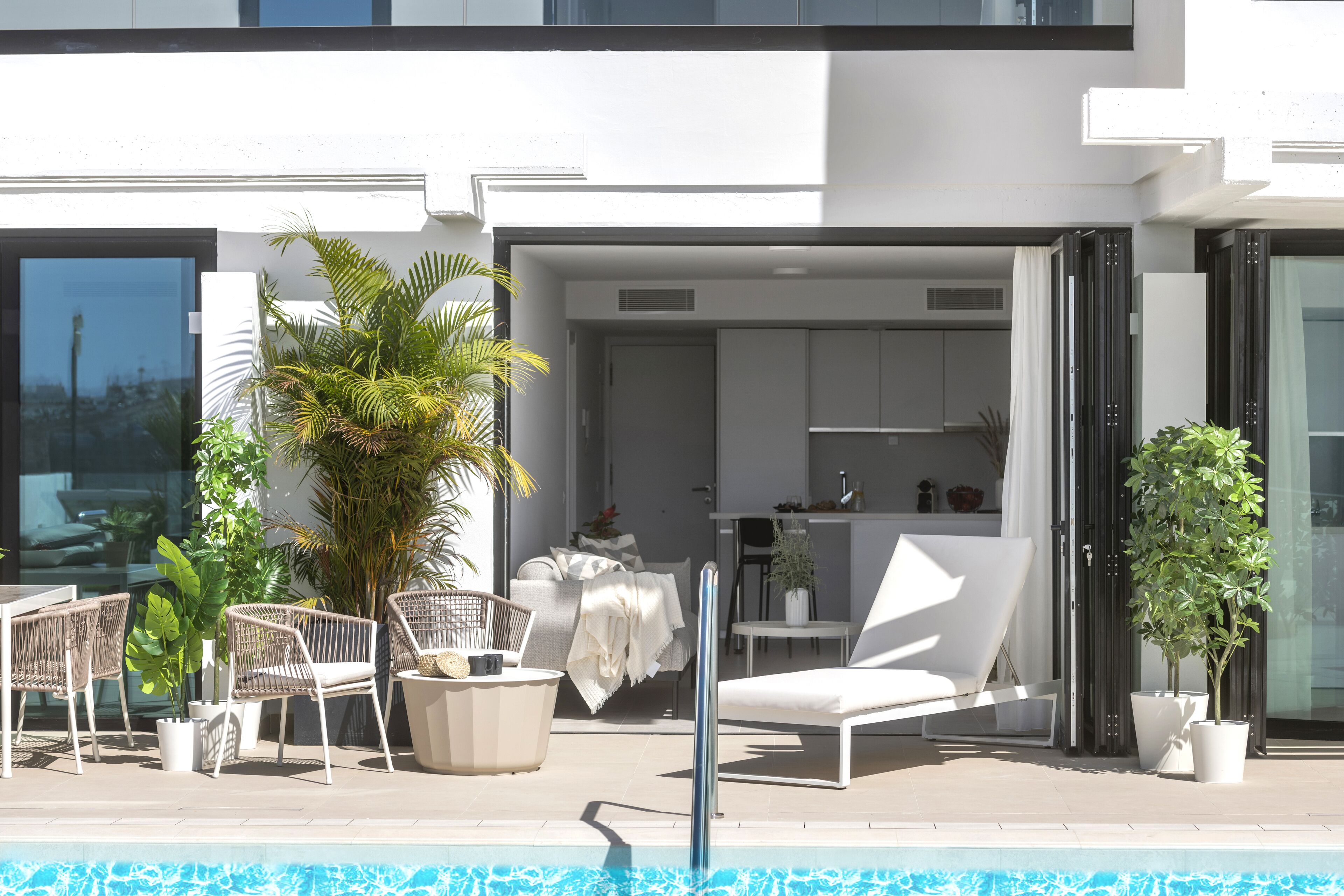 A modern outdoor pool area with lounge chairs, potted plants, and a kitchen visible through an open door.