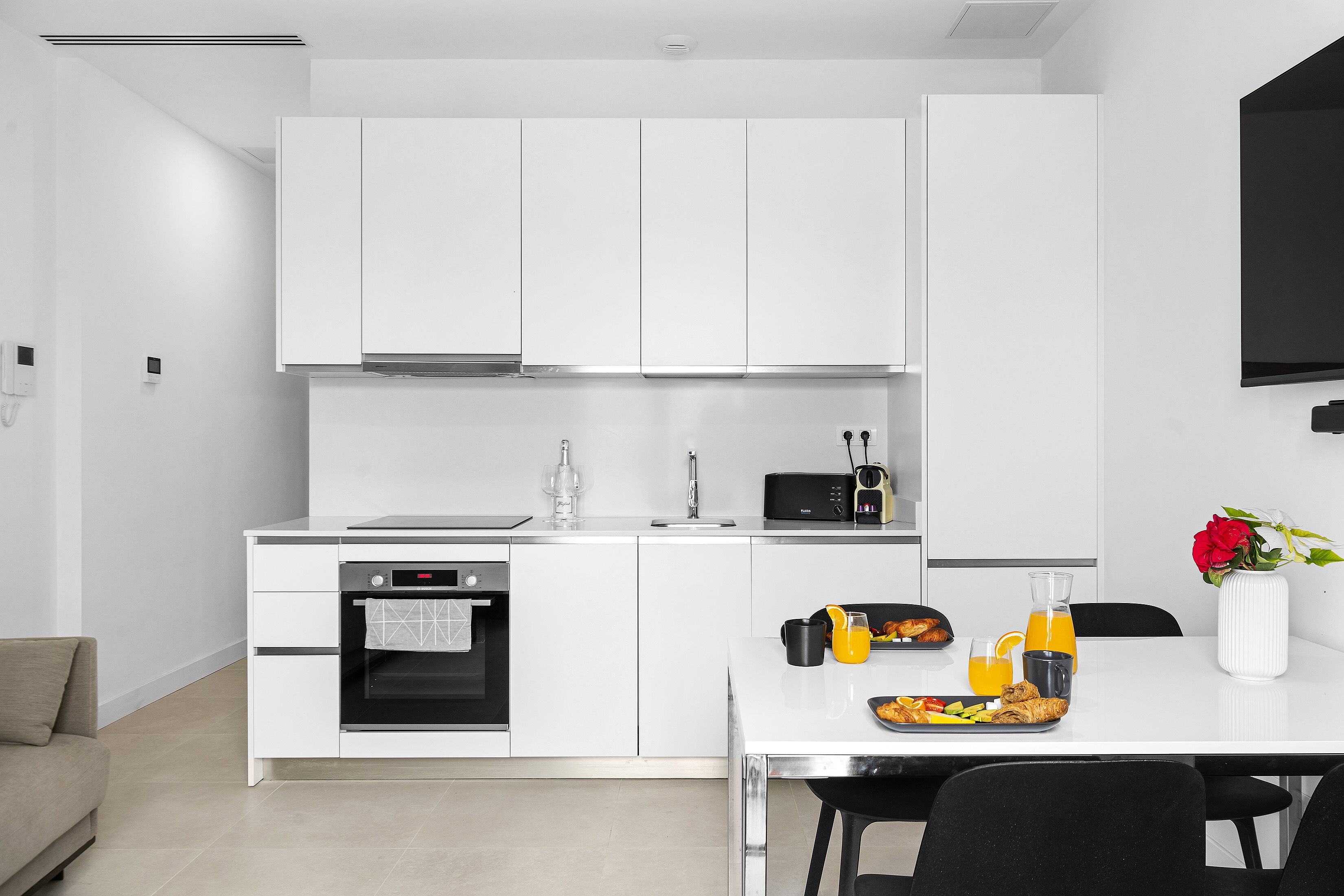A modern kitchen with white cabinets, a built-in oven, a dining table with chairs, and a breakfast tray.
