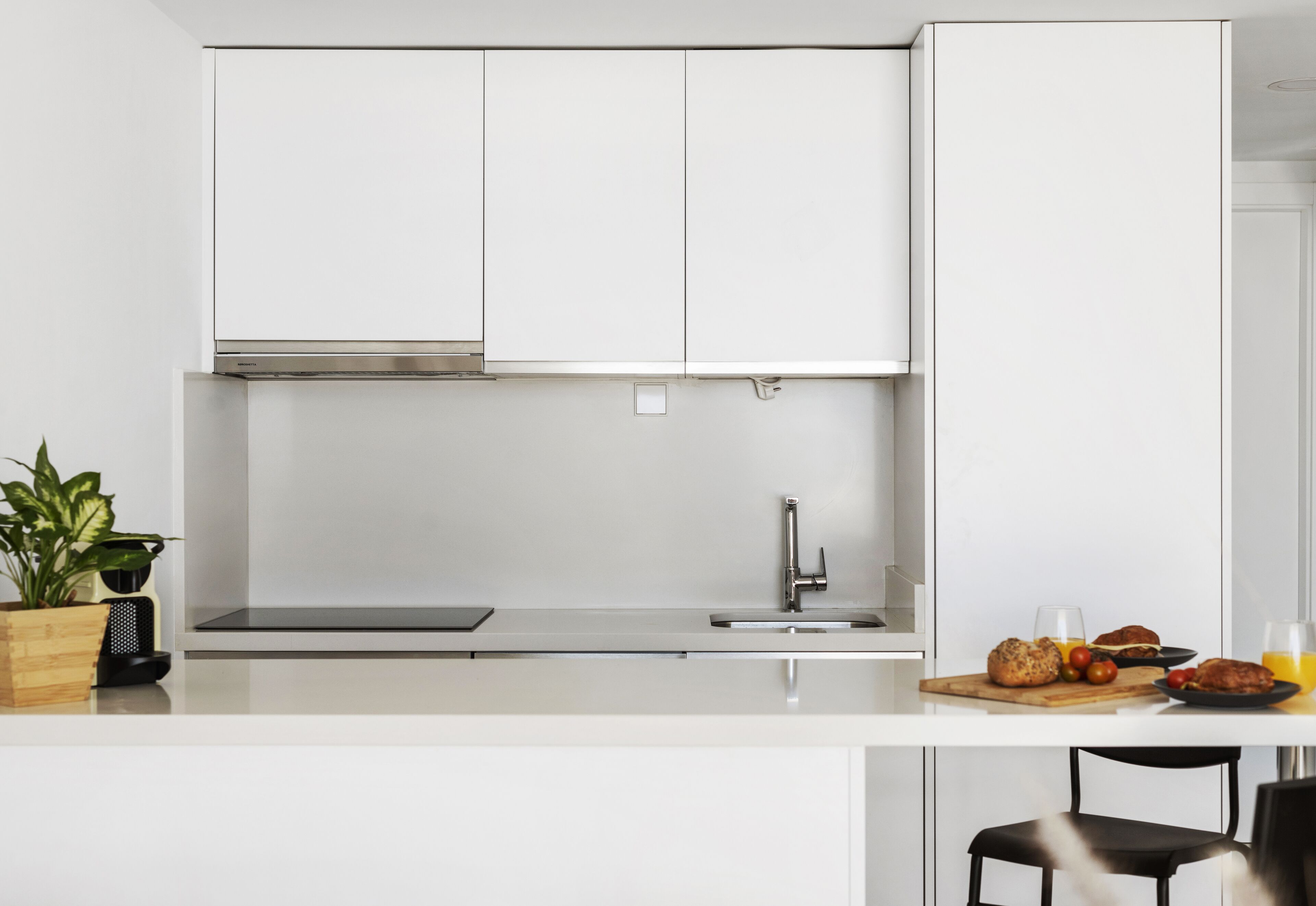 Una cocina moderna con armarios blancos, una campana extractora de acero inoxidable, un fregadero y una zona de desayuno con pan, tomates y jugo.