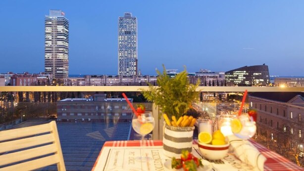 A table with a menu, drinks, and a plant on a balcony overlooking a cityscape at dusk. - ENJOYBCN MARINA APARTMENT (Barcelona)