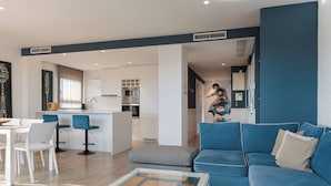 A modern living room with a blue sofa, a glass coffee table, and a dining area with white chairs. - ENJOYBCN MARINA APARTMENT (Barcelona)