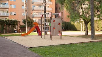 Un parque infantil con tobogán y estructuras para escalar, rodeado de una zona pavimentada y árboles, con un edificio al fondo.