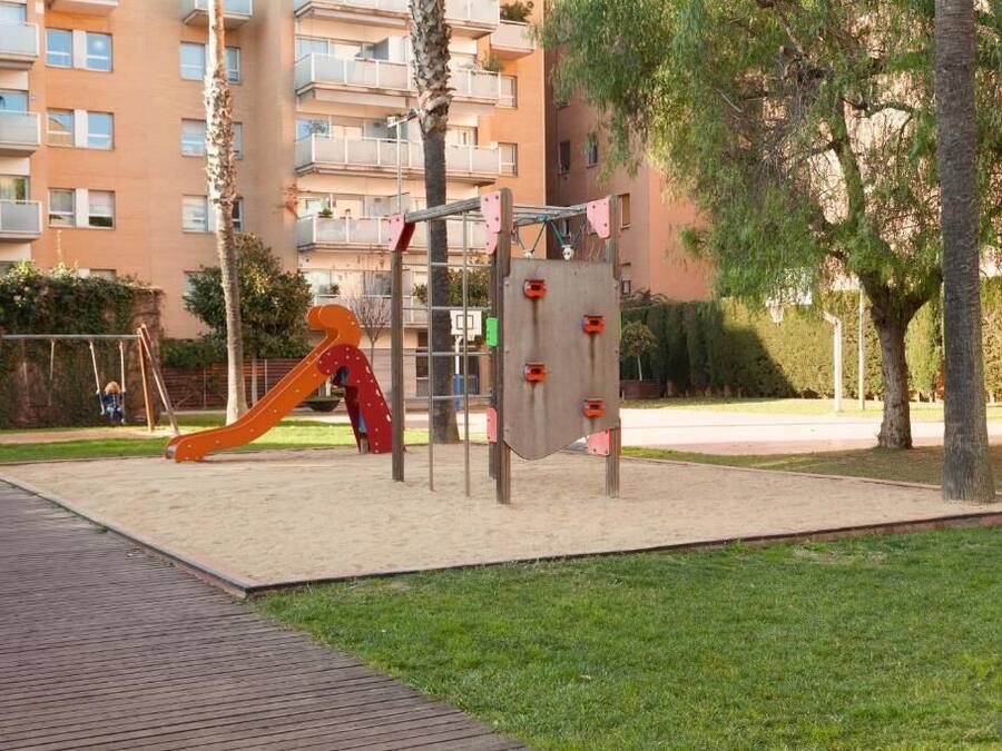 Un parque infantil con tobogán y estructuras para escalar, rodeado de una zona pavimentada y árboles, con un edificio al fondo.