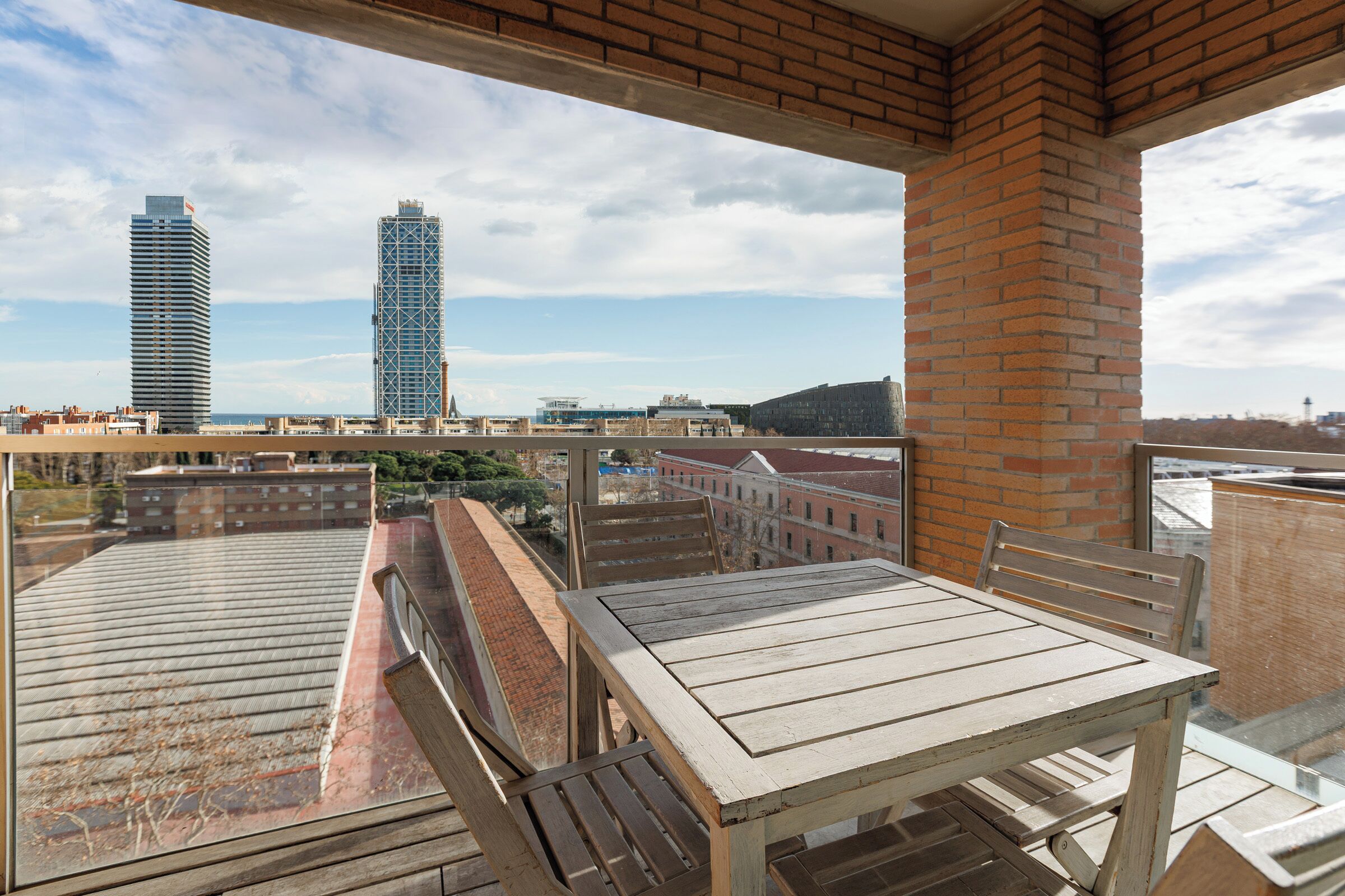 A balcony with a wooden table and chairs, overlooking a cityscape with buildings and a clear sky.